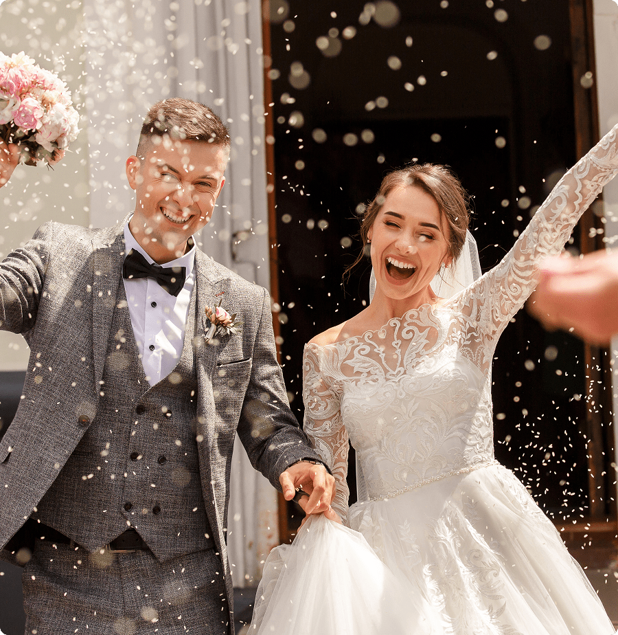GettyImages-1190043570 A joyful bride and groom celebrate outside with confetti. The bride holds pink flowers and wears a lace gown, while the groom is in a gray suit and bow tie.